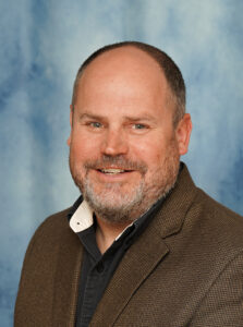 "Portrait of a smiling middle-aged man in a brown blazer, against a blue backdrop."